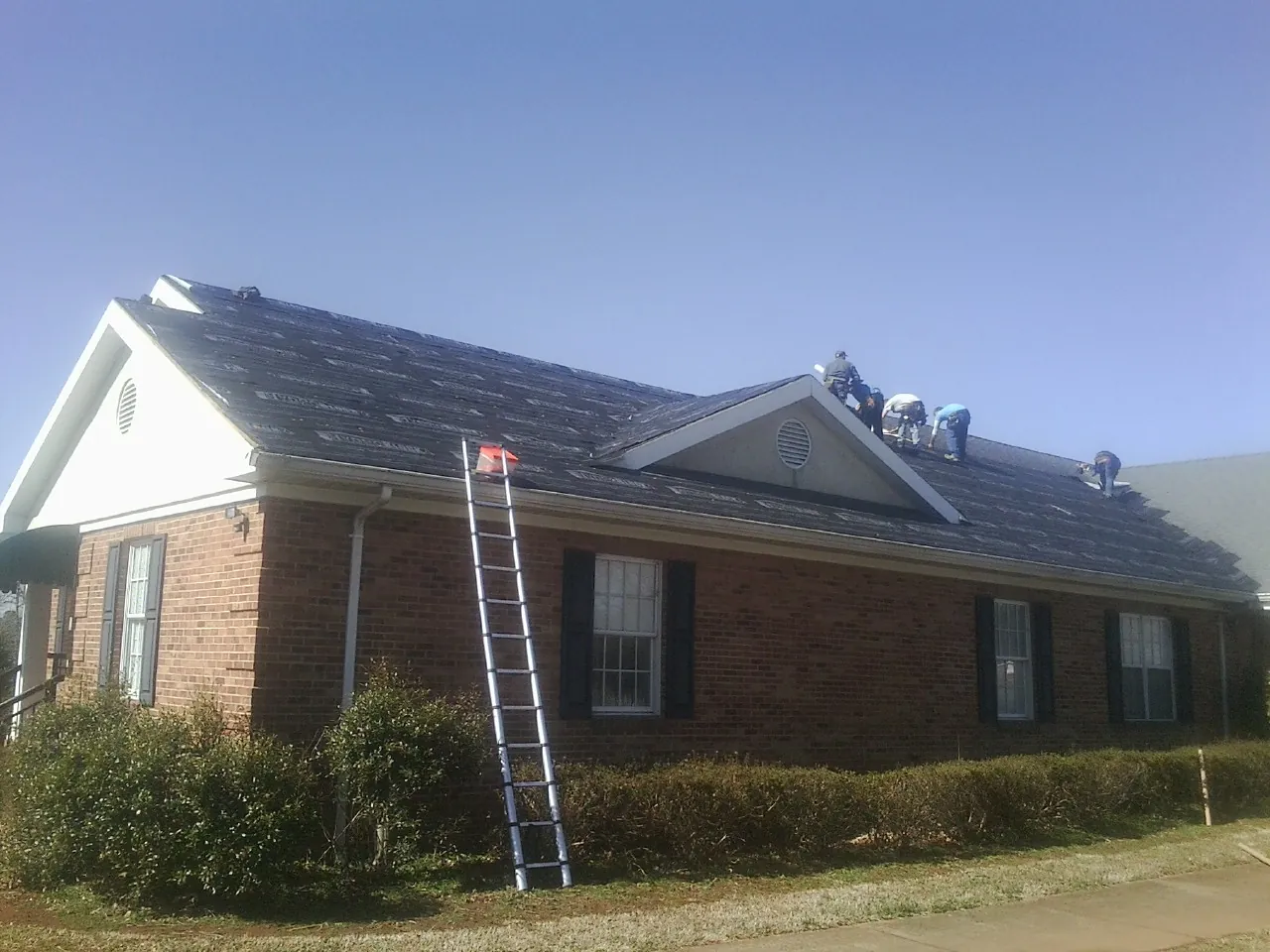 Crew tearing shingles off a brick ranch roof with a ladder set — Metal Roof Repair work by our crew in Atkinson