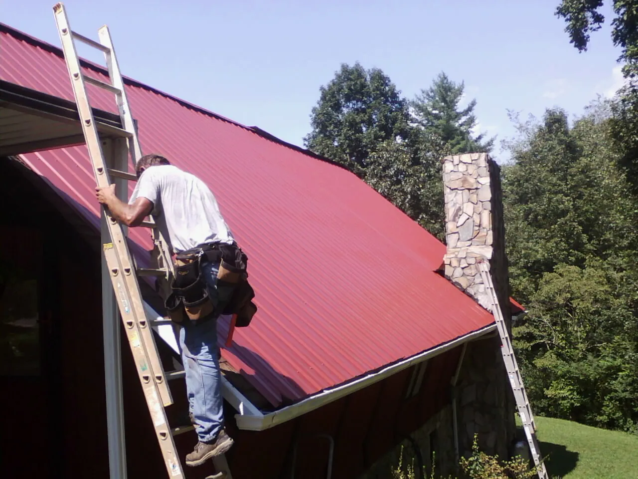 Roofer on a ladder beside a red metal roof and stone chimney — Soffit & Fascia Repair by Roofing Brothers in Atkinson